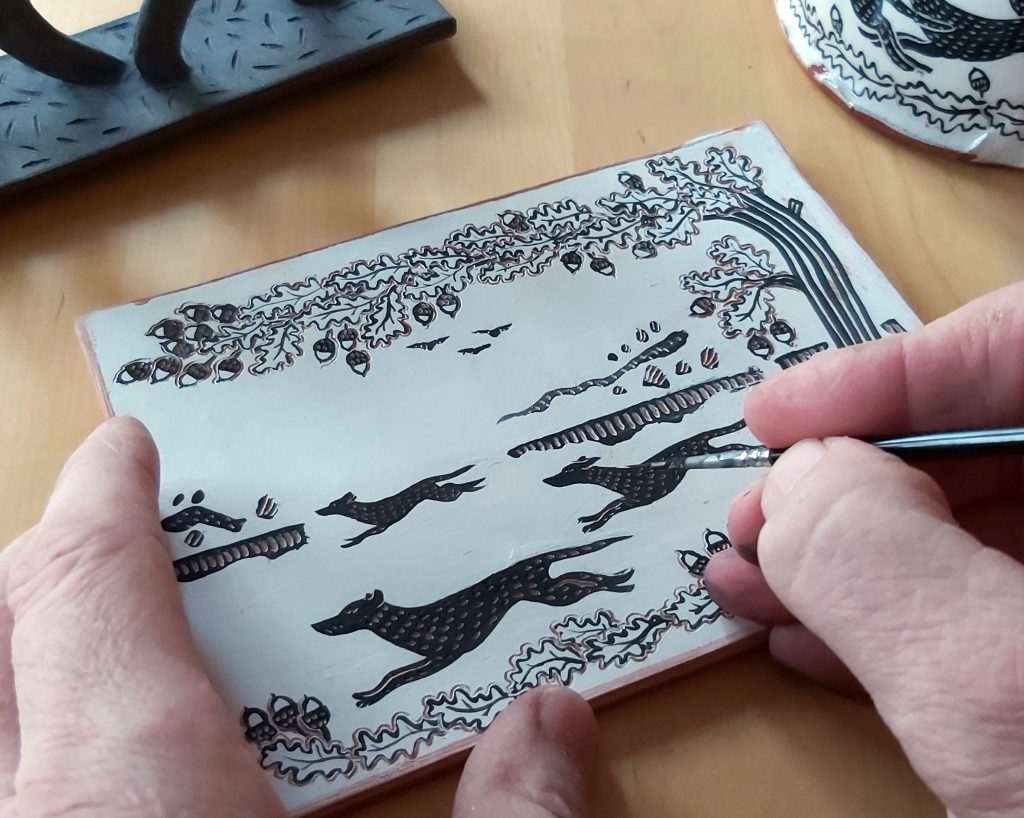 Image shows a close up of Simon Olley's hands illustrating an image of three black dogs running through wooded countryside onto a flat ceramic postcard, black image on white clay.